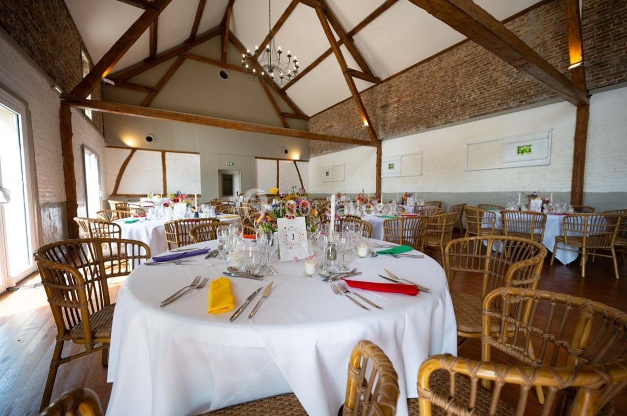 Salle de réception lumineuse avec tables rondes dressées, chaises en rotin et poutres apparentes au plafond.