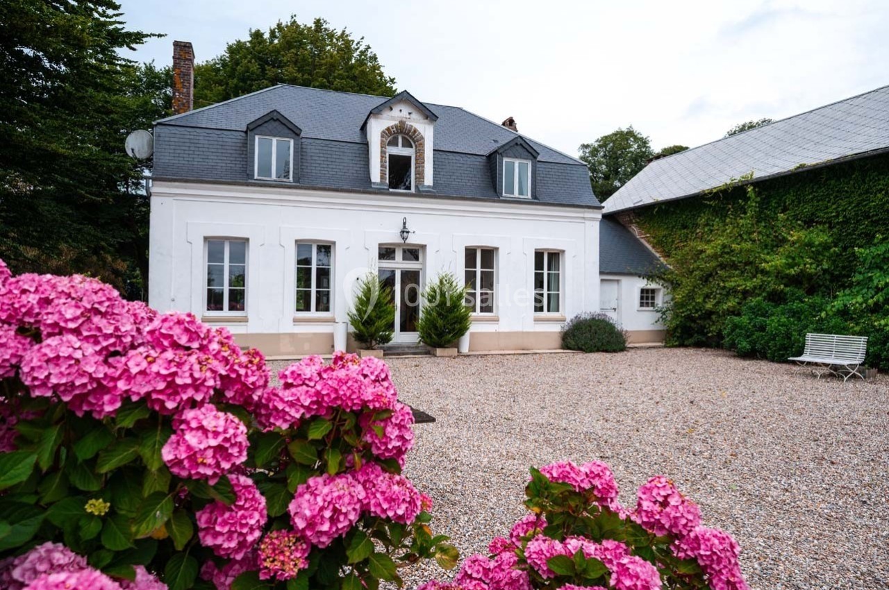 Façade d'une maison blanche avec toit en ardoise, devant une cour gravillonnée et des hortensias roses au premier plan.