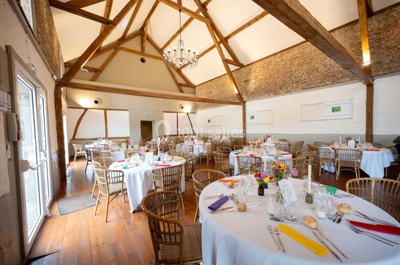 Salle de réception lumineuse avec tables rondes dressées, chaises en osier et poutres apparentes au plafond.