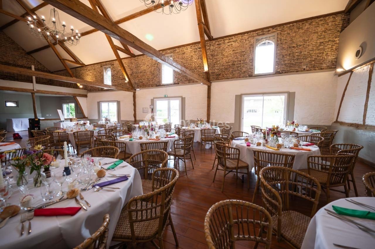Salle de réception lumineuse avec murs en briques, poutres apparentes et tables rondes décorées pour un événement.