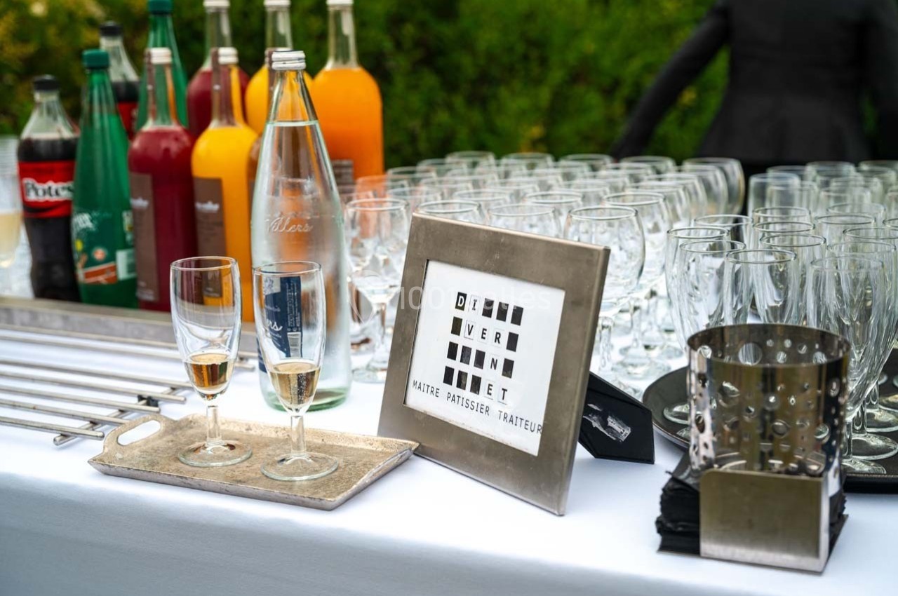 Buffet avec bouteilles de boissons, verres vides, deux verres remplis et un cadre affichant le nom d’un traiteur.