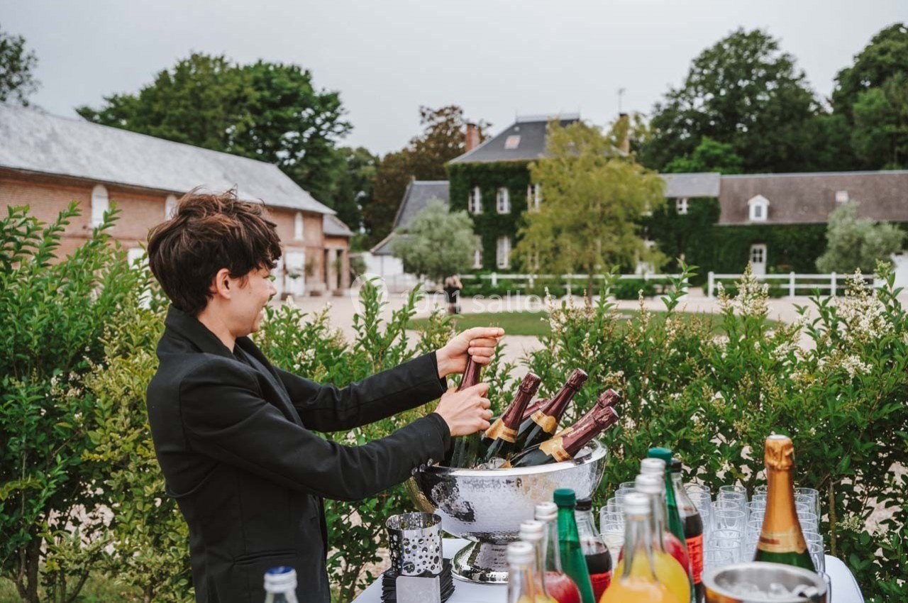 Une personne ouvre une bouteille de champagne près d'une table garnie de boissons, dans un jardin devant des bâtiments.
