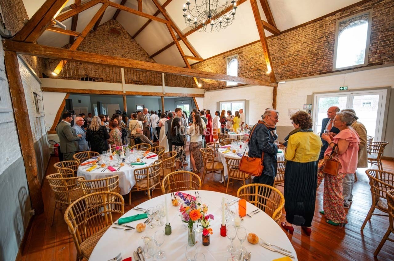 Salle de réception avec des invités debout et assis autour de tables décorées, sous une charpente en bois apparente.