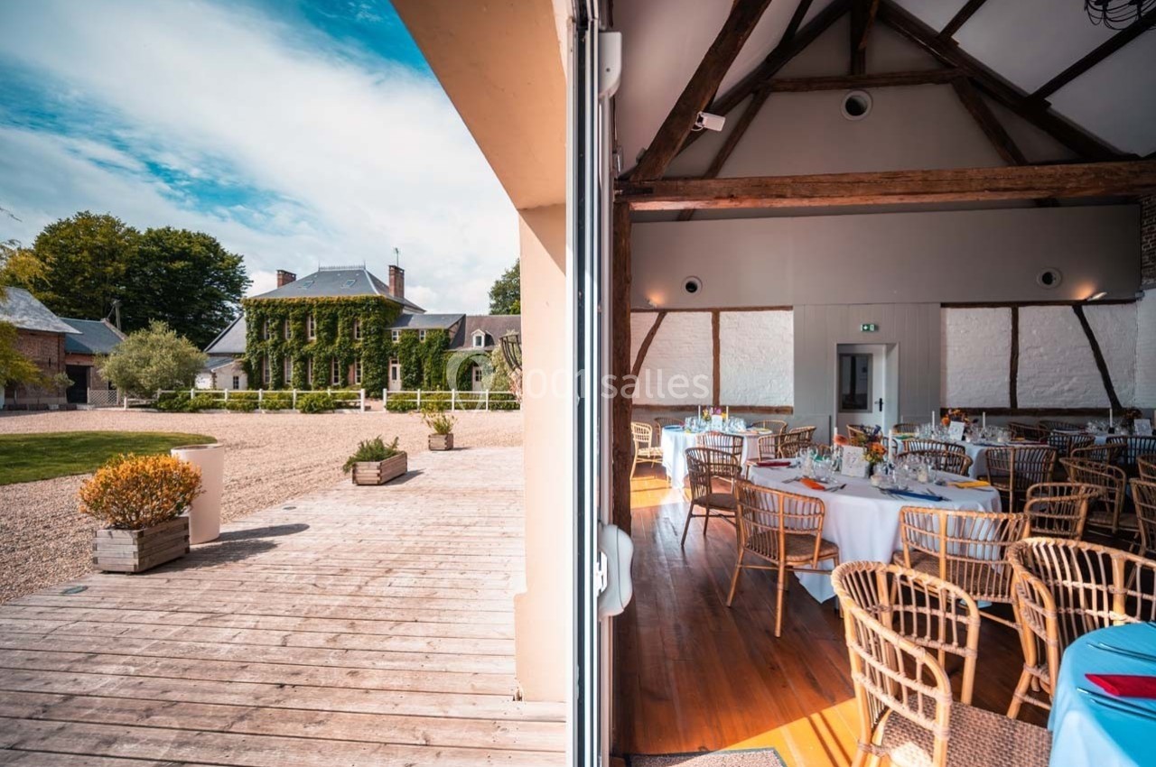 Vue d'une salle de réception décorée et d'une terrasse en bois donnant sur un jardin et une maison en pierre.