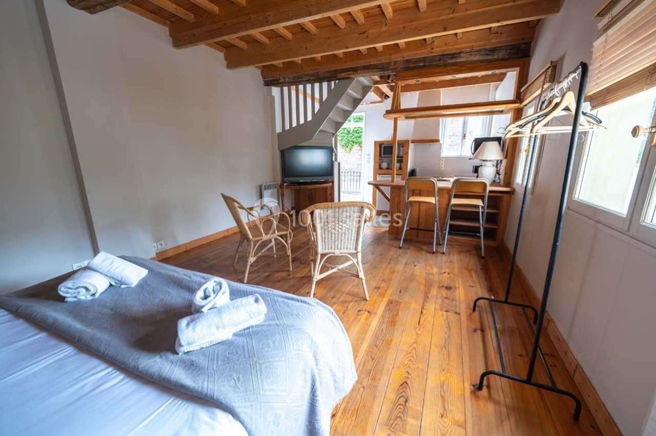 Chambre lumineuse avec lit double, coin salon en rotin, bureau en bois, télévision et parquet en bois clair.