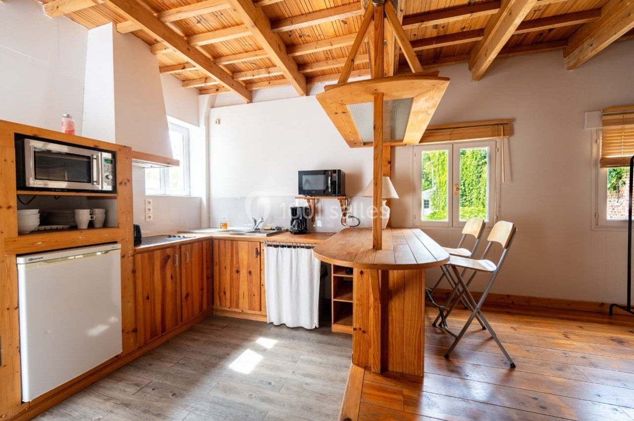 Cuisine en bois avec plafond à poutres apparentes, équipée d'un micro-ondes, réfrigérateur, évier et coin repas.