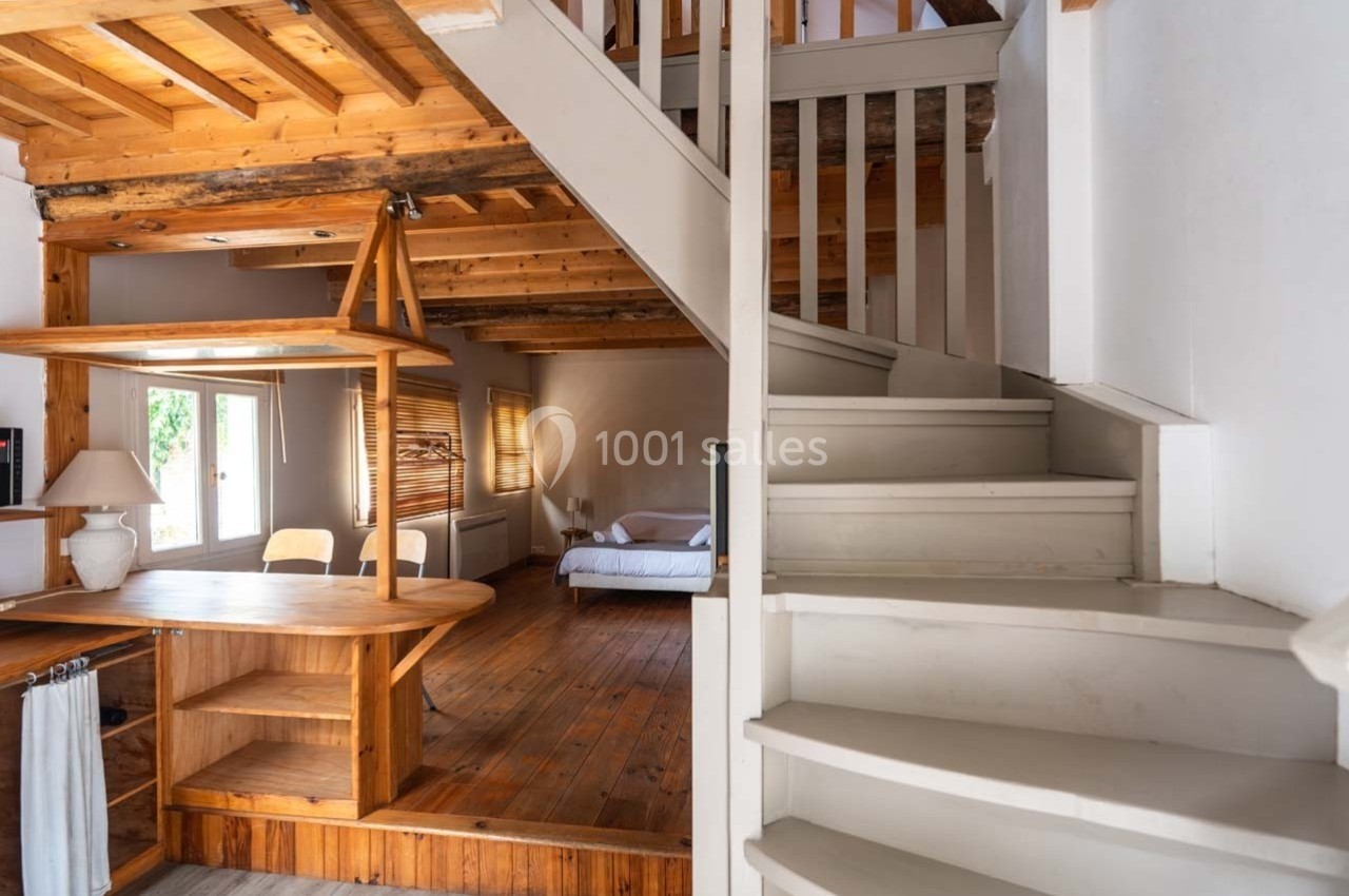Escalier en bois blanc menant à un étage, avec un salon en arrière-plan doté de meubles en bois et d'une grande fenêtre.