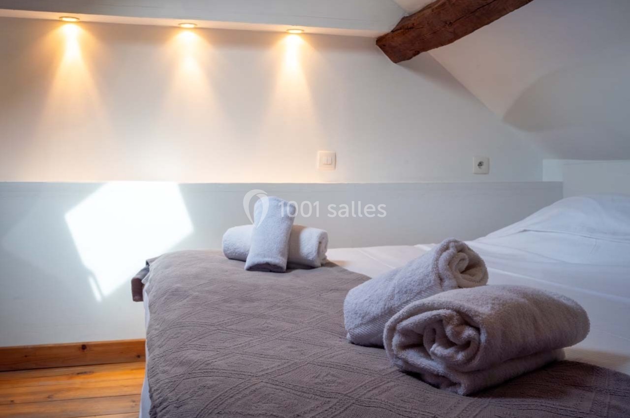 Lit double avec couverture grise, serviettes roulées et lumière douce sous un plafond mansardé avec poutre apparente.