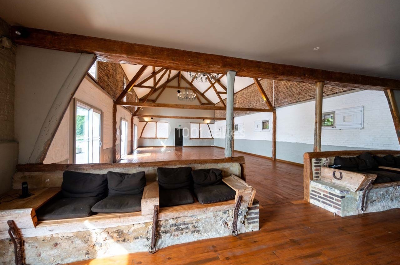Salle spacieuse avec poutres apparentes, murs en briques blanches et parquet en bois, éclairée par des fenêtres latérales.