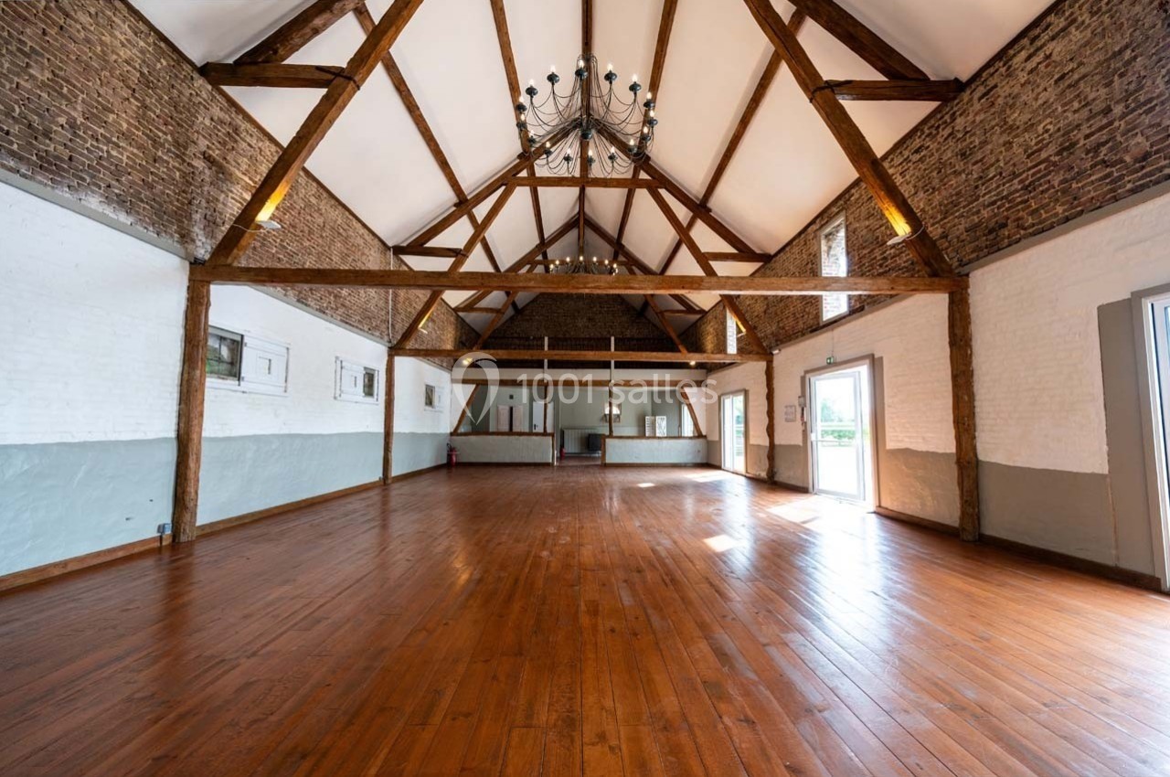 Grande salle lumineuse avec parquet en bois, murs en briques et poutres apparentes, éclairée par un lustre central.