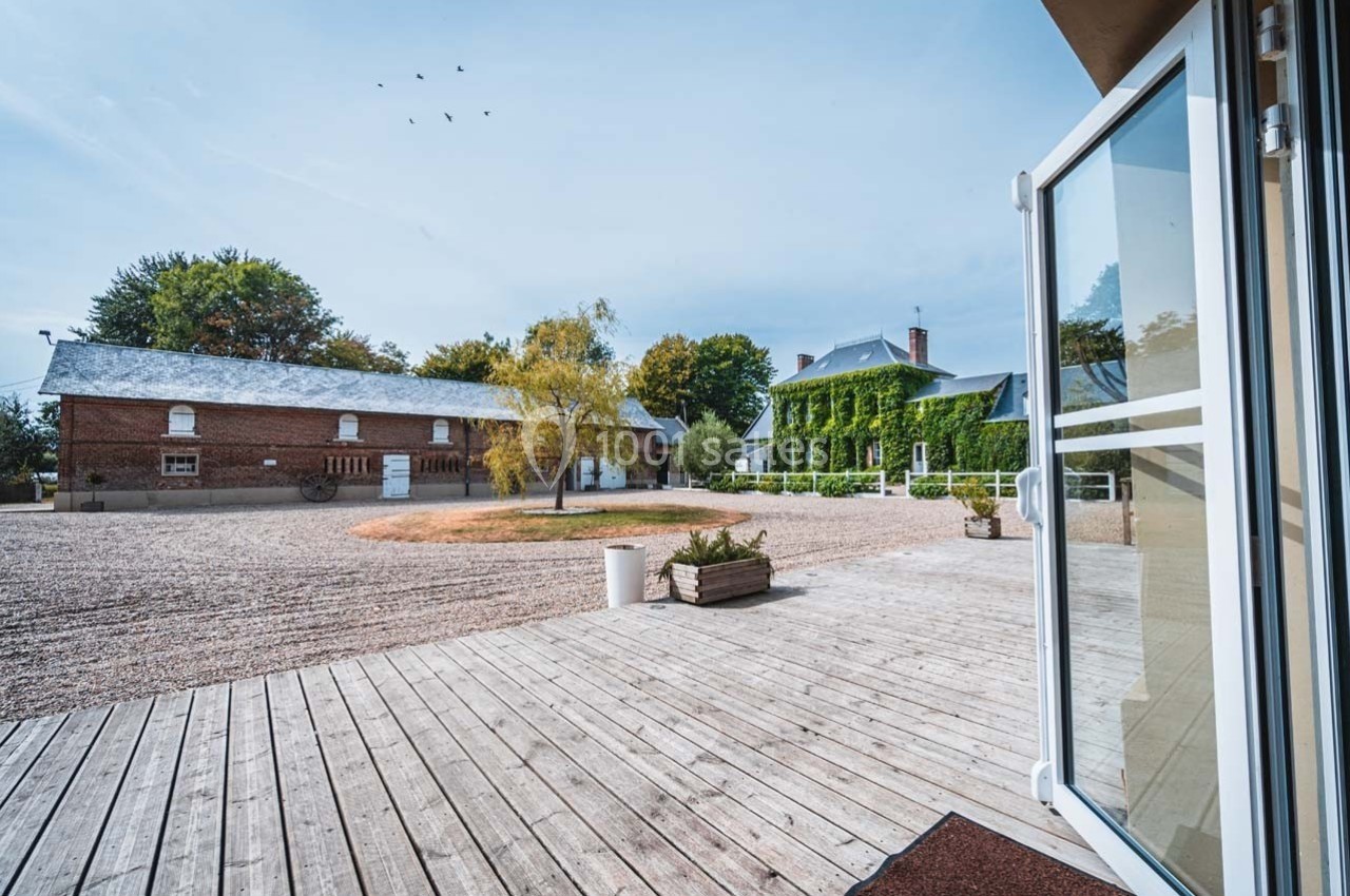 Vue sur une cour avec bâtiments en briques et maison couverte de végétation, depuis une terrasse en bois.