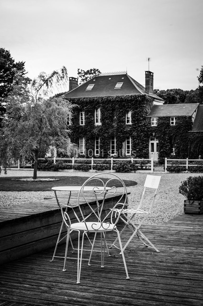 Chaises et table en fer forgé sur une terrasse en bois, avec une maison couverte de lierre en arrière-plan.