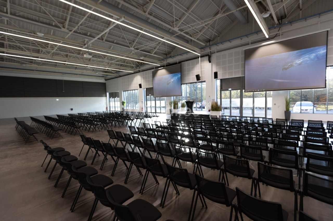 Salle de conférence moderne avec rangées de chaises noires, écrans géants et grandes baies vitrées.