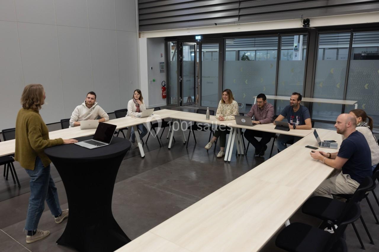Une personne présente devant un groupe de collègues assis autour d'une table en U dans une salle de réunion moderne.