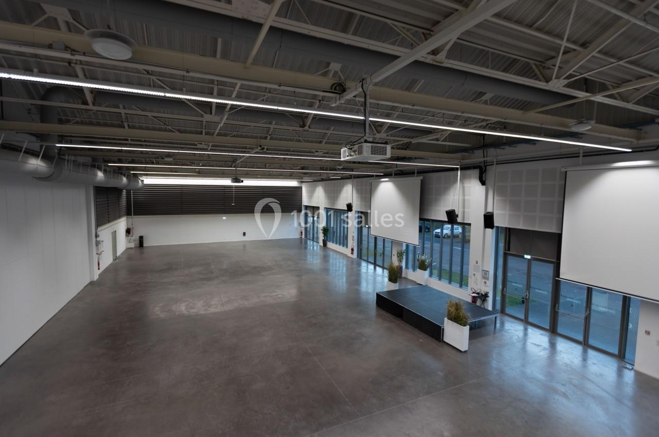 Salle vide avec sol en béton, grandes baies vitrées, murs blancs et plafond industriel avec éclairage suspendu.