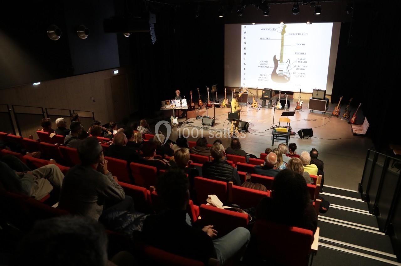 Conférence dans une salle avec public, présentation sur écran et plusieurs guitares exposées sur scène.