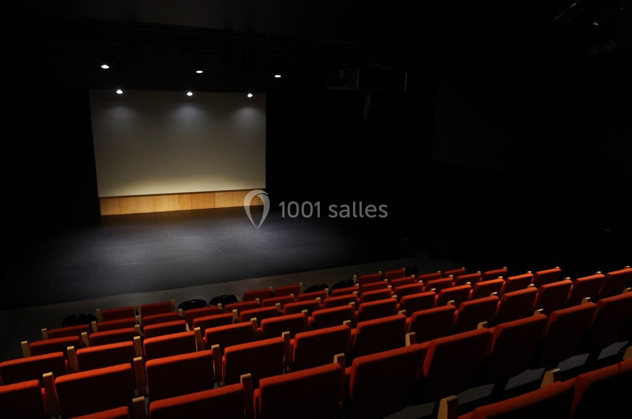 Salle de spectacle vide avec des rangées de sièges rouges face à une scène éclairée et un écran blanc.