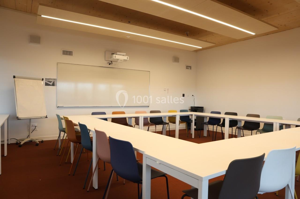 Salle de réunion lumineuse avec tables disposées en U, chaises colorées, tableau blanc et projecteur au mur.