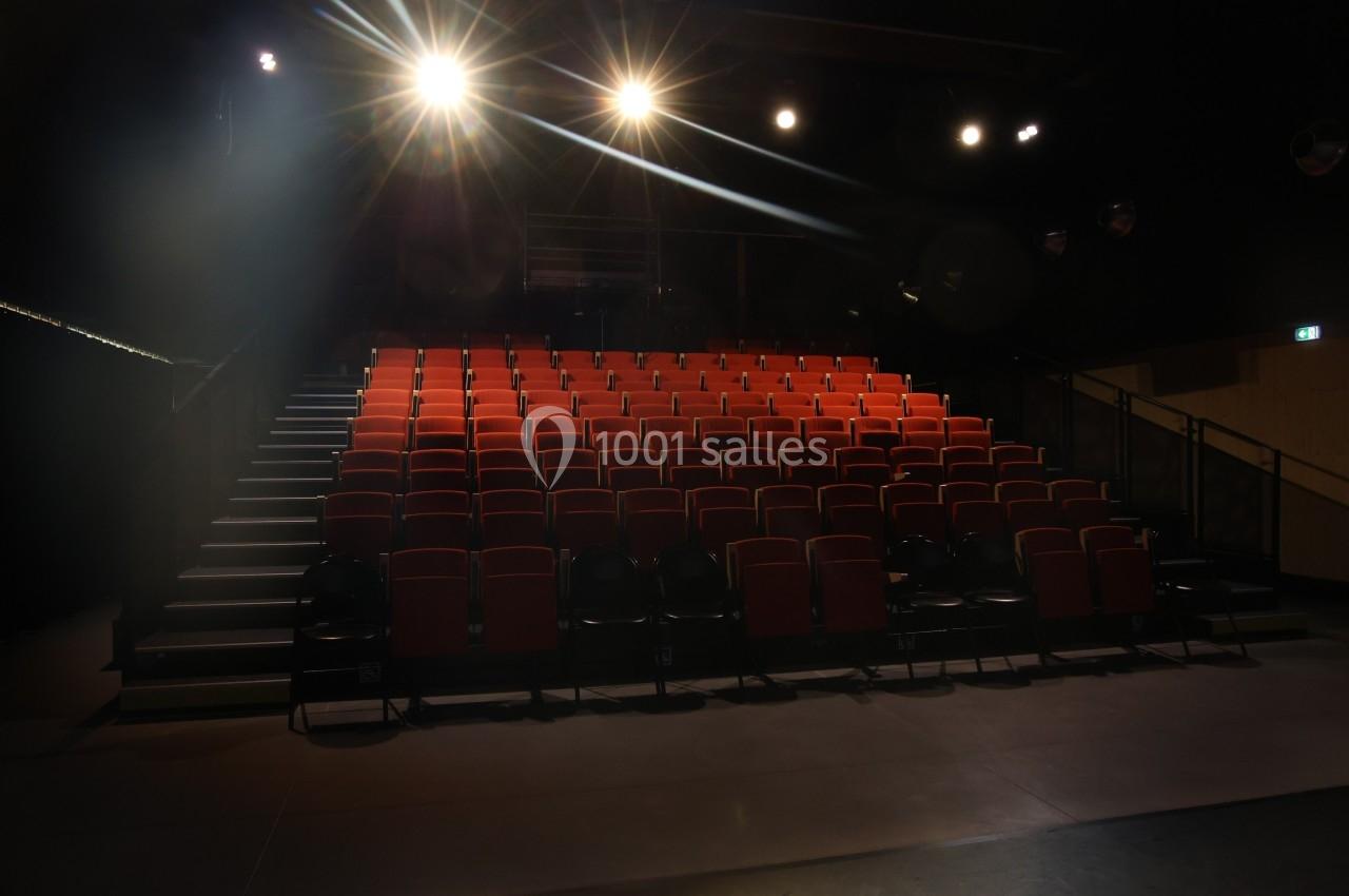 Salle de spectacle vide avec des rangées de sièges rouges et des projecteurs allumés éclairant la scène.