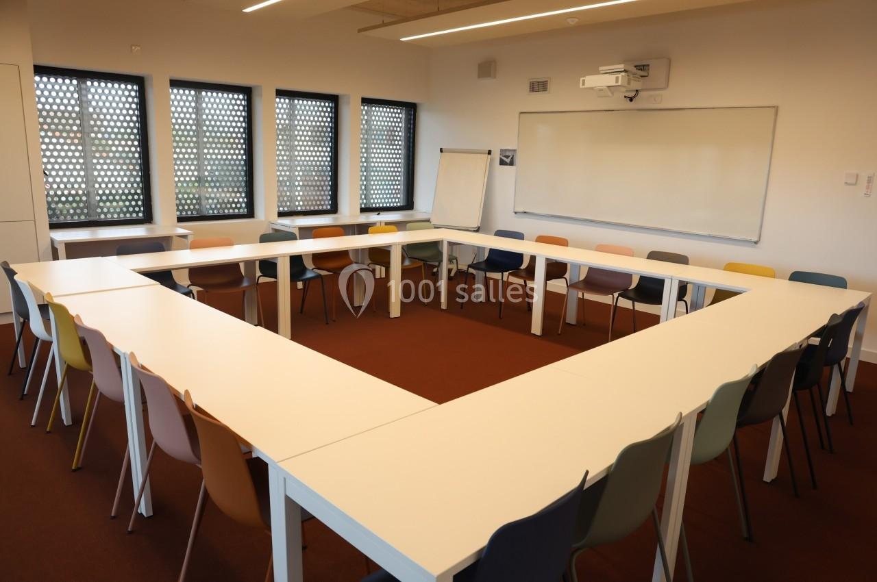 Salle de réunion avec tables disposées en U, chaises colorées, tableau blanc et paperboard près des fenêtres.