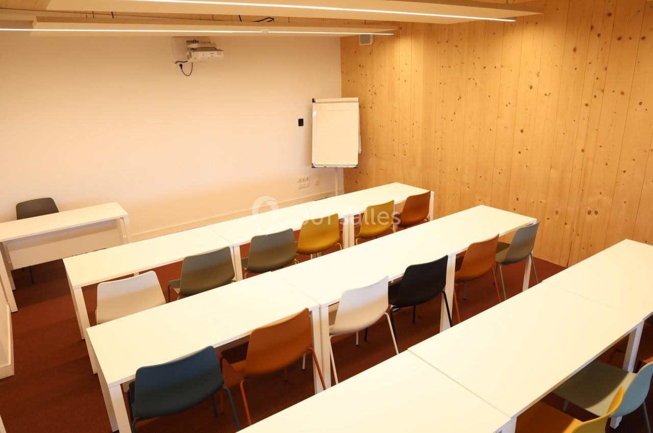 Salle de formation lumineuse avec des tables blanches alignées, des chaises colorées et un tableau de présentation.