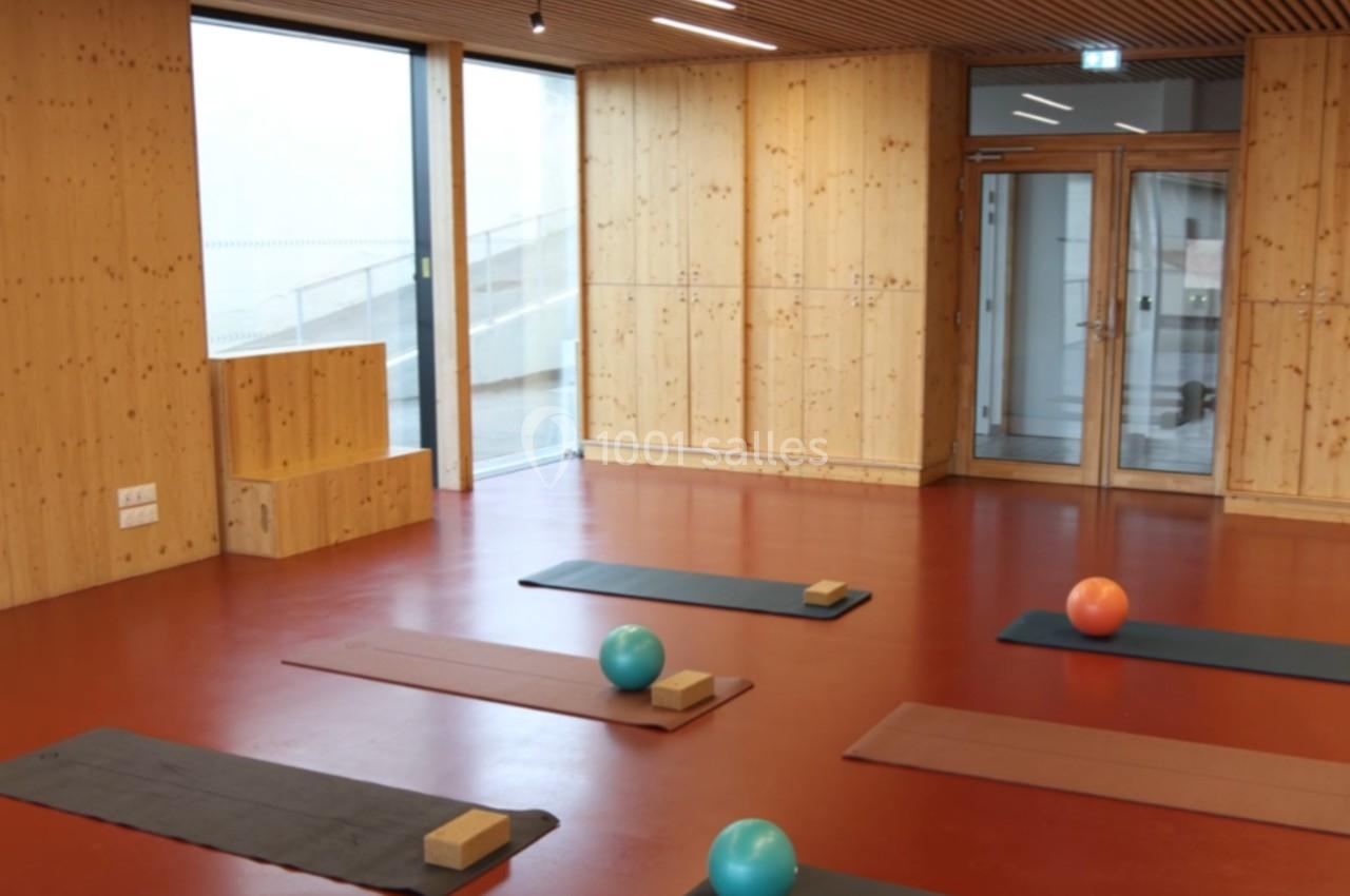 Salle lumineuse avec sol rouge, murs en bois, tapis de yoga, ballons et briques disposés pour une séance.