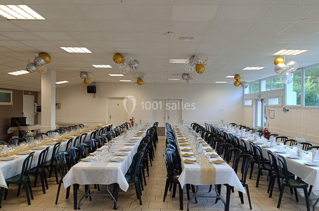Salle décorée avec des tables alignées, nappes blanches et chemins de table dorés, prête pour un événement festif.