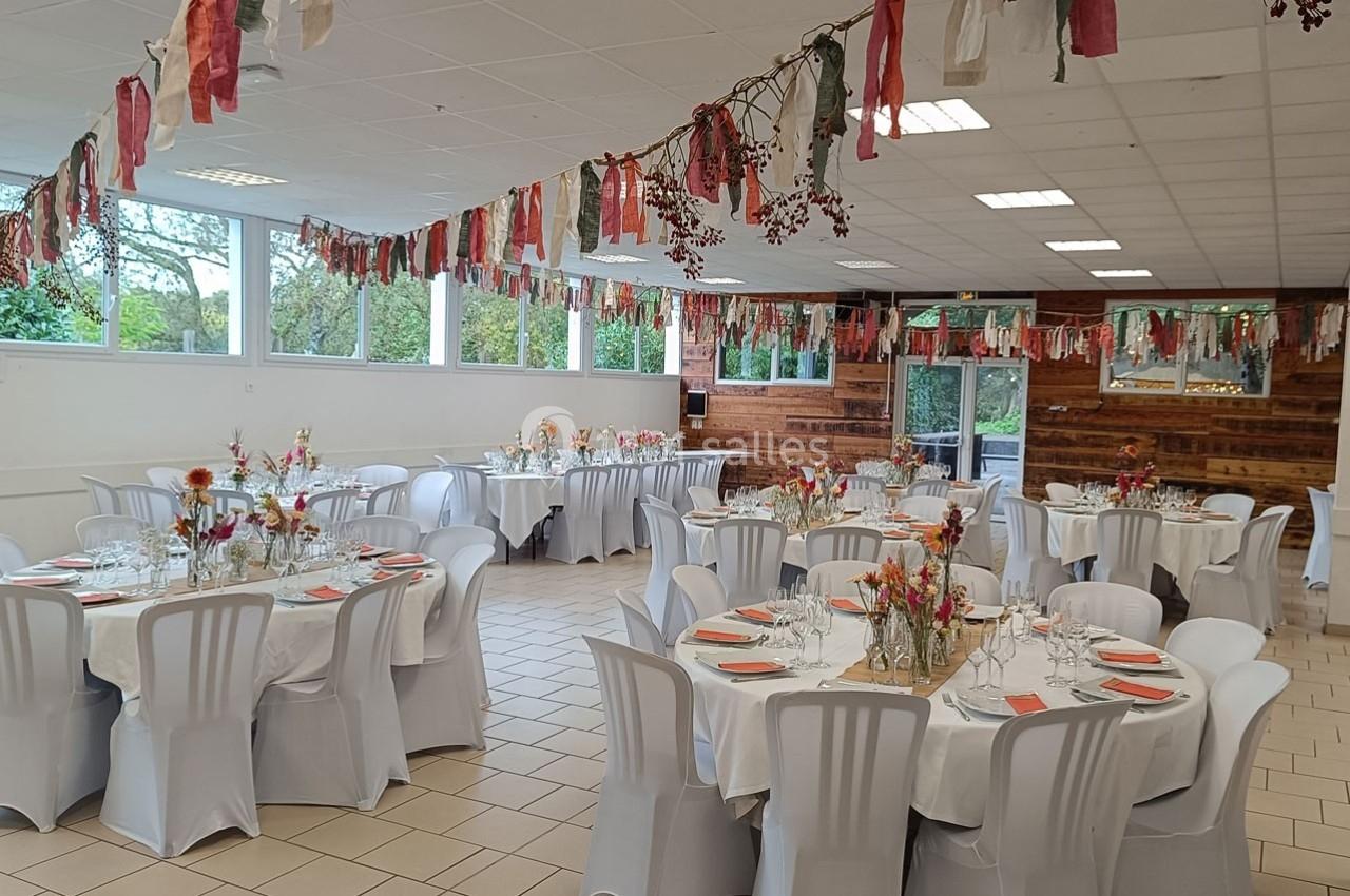 Salle de réception décorée avec des tables rondes dressées, des chaises blanches et des guirlandes suspendues colorées.