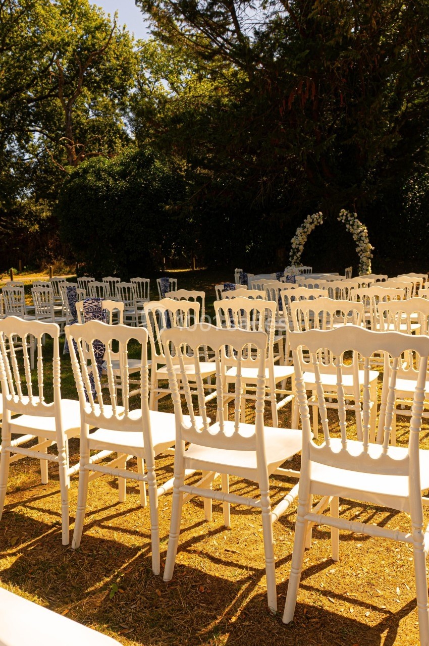 Chaises blanches alignées en extérieur devant une arche fleurie, sous des arbres par une journée ensoleillée.
