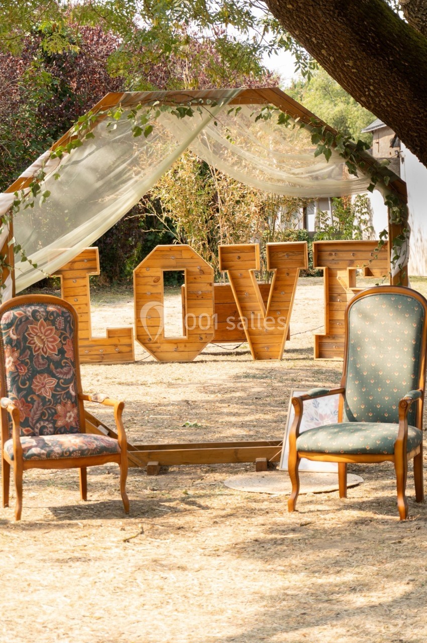 Deux fauteuils anciens devant une structure en bois formant le mot ’LOVE’, sous un arbre dans un décor champêtre.