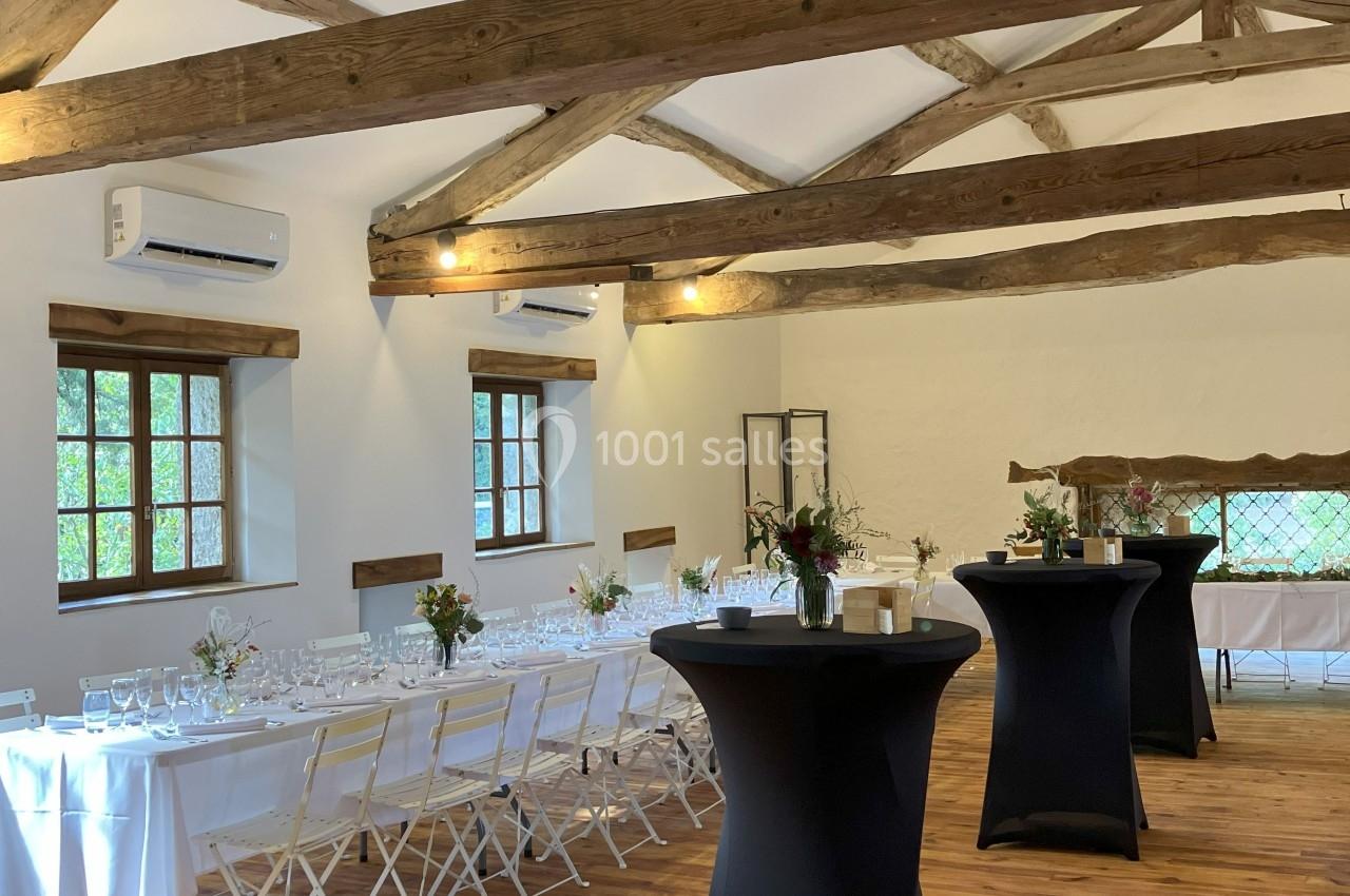 Salle lumineuse avec poutres apparentes, tables dressées pour un repas et tables hautes noires sur parquet en bois.