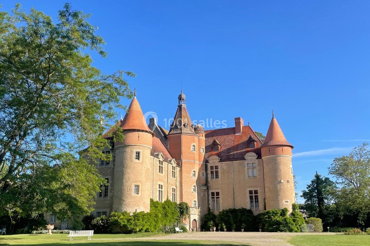 Château en pierre avec tours et toits rouges, entouré de pelouse et d'arbres sous un ciel bleu dégagé.