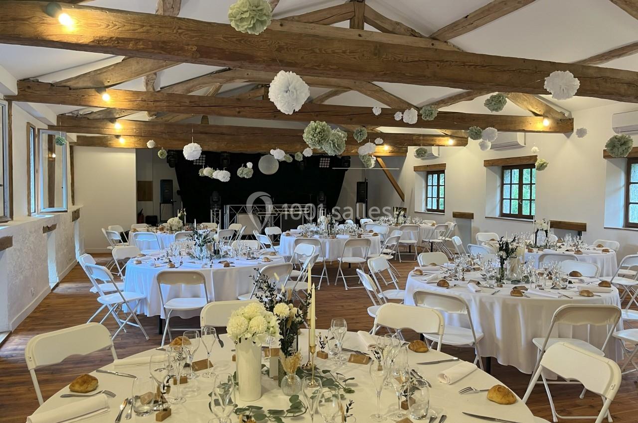 Salle de réception décorée pour un événement, avec tables rondes dressées, chaises blanches et guirlandes suspendues.