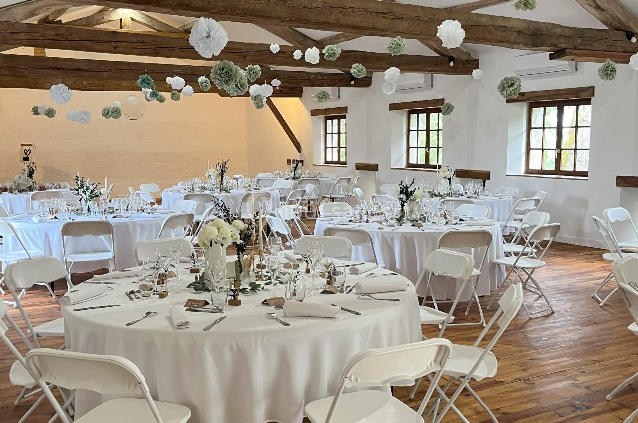 Salle de réception décorée avec des tables rondes, nappes blanches, chaises pliantes et suspensions en papier.
