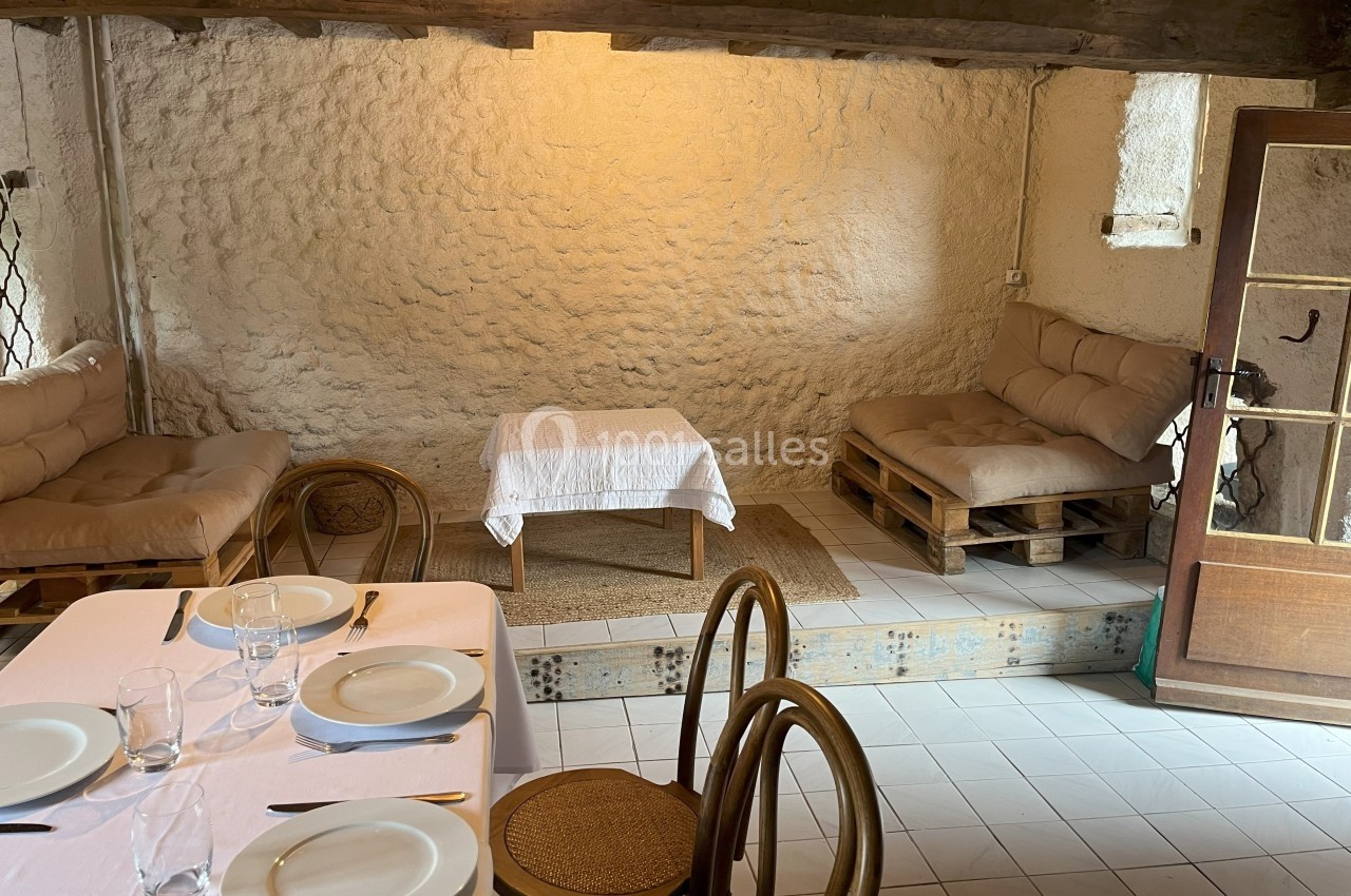 Salle rustique avec murs en pierre, table dressée au premier plan et coin salon avec palettes en arrière-plan.