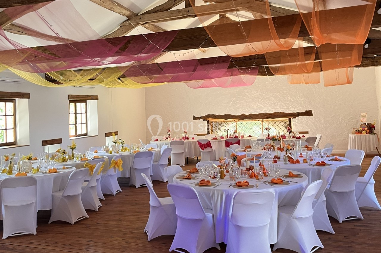 Salle de réception décorée avec des tables rondes dressées, nappes blanches et voilages colorés suspendus au plafond.