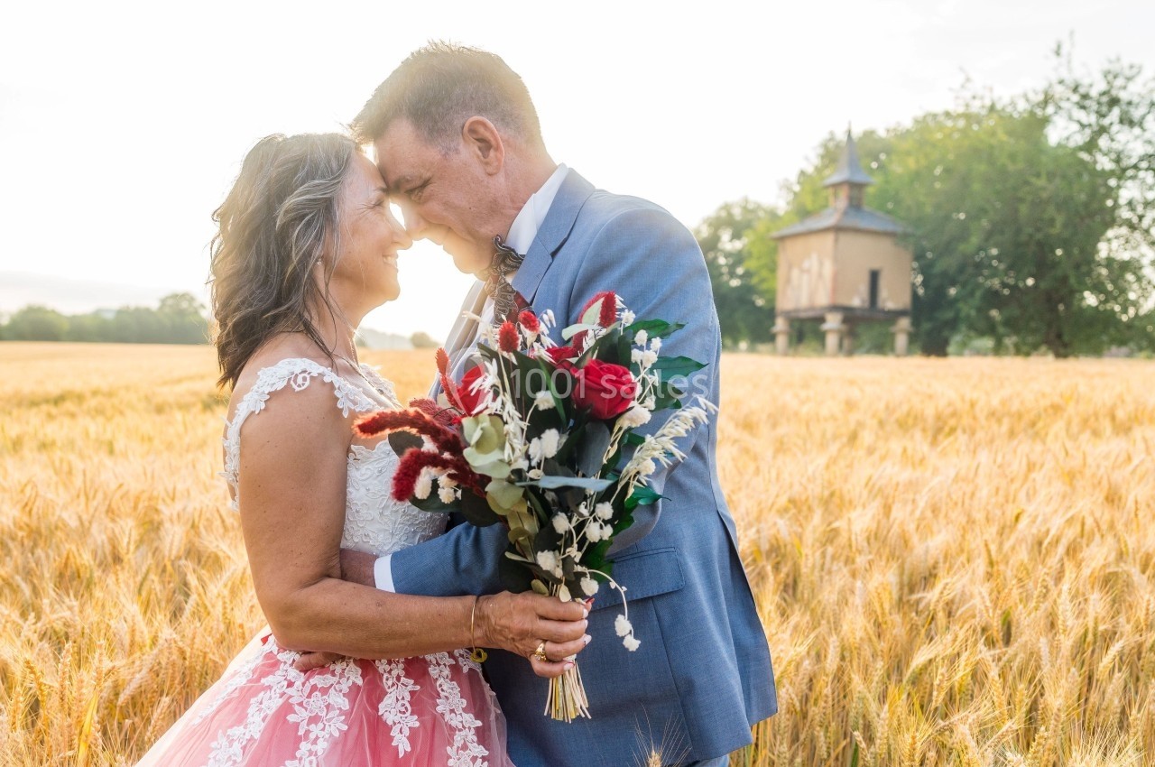 Un couple souriant s'enlace dans un champ de blé au coucher du soleil, tenant un bouquet de fleurs.