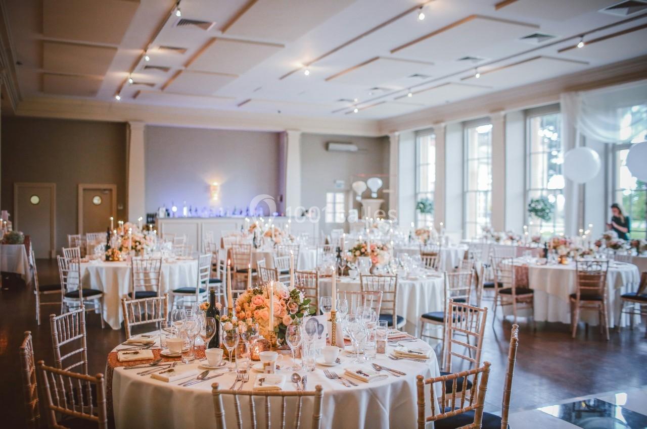 Salle de réception élégante avec tables rondes décorées de fleurs et vaisselle, éclairée par de grandes fenêtres.