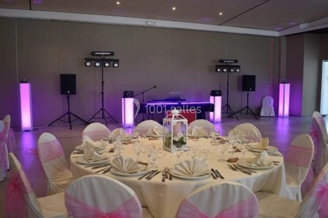 Salle de réception décorée avec une table ronde dressée, chaises ornées de nœuds roses et un espace avec matériel de…