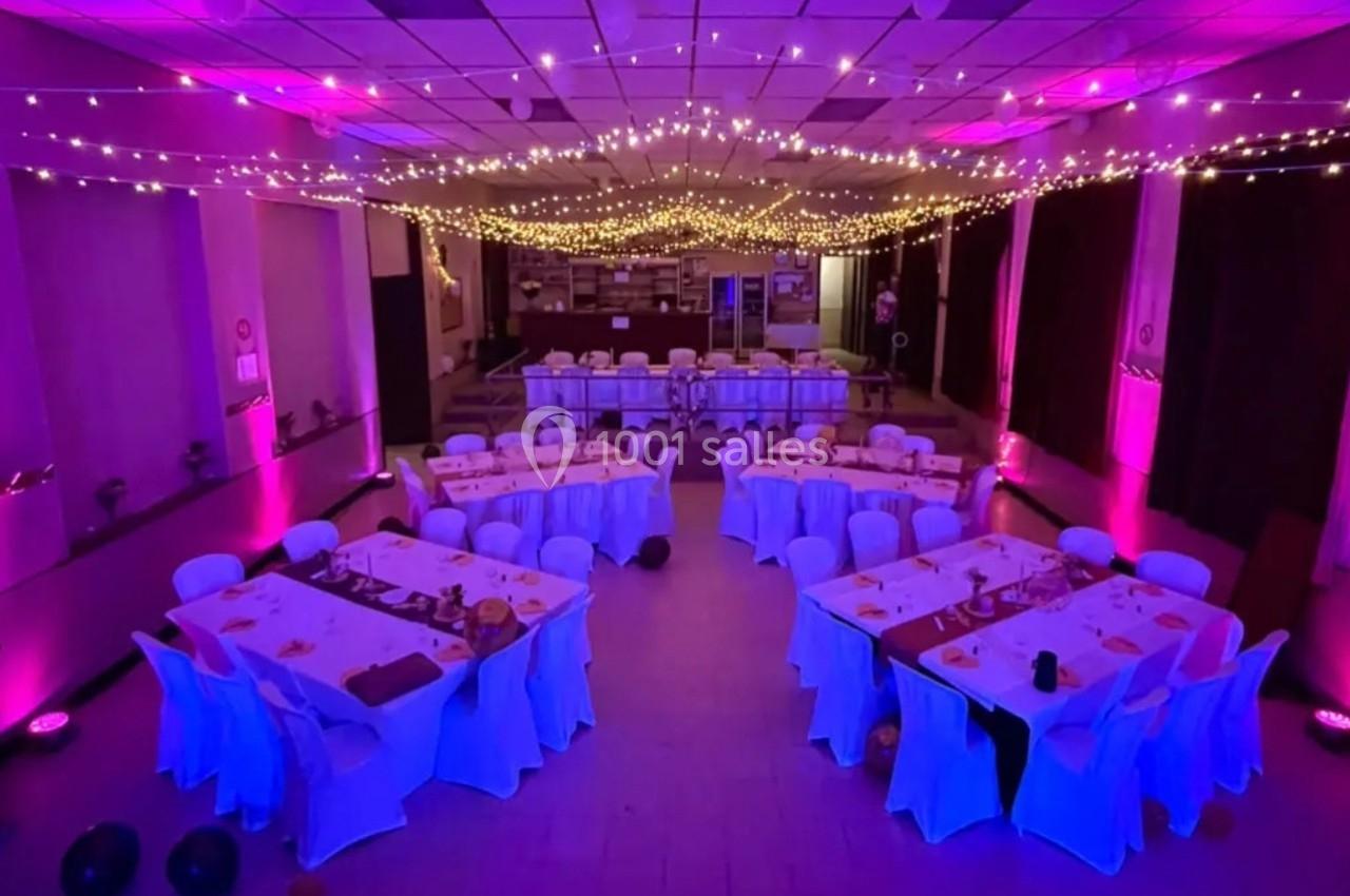 Salle de réception décorée avec des guirlandes lumineuses au plafond, tables dressées et éclairage rose.