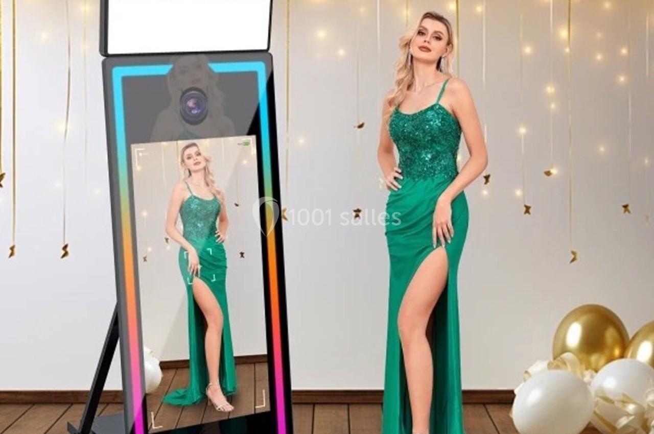 Une femme en robe verte élégante posant devant un miroir interactif, avec des ballons dorés en arrière-plan.