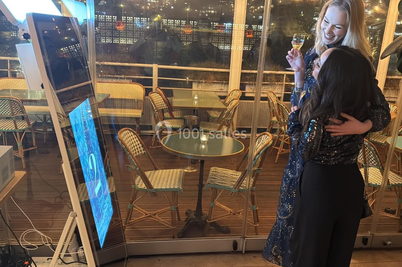 Deux femmes souriantes se tiennent près d'une borne photo dans un espace intérieur avec des tables et des chaises en arrière…