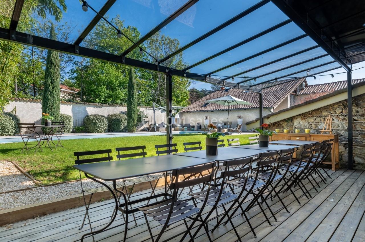 Terrasse en bois avec tables et chaises sous une pergola vitrée, donnant sur un jardin avec piscine et transats.