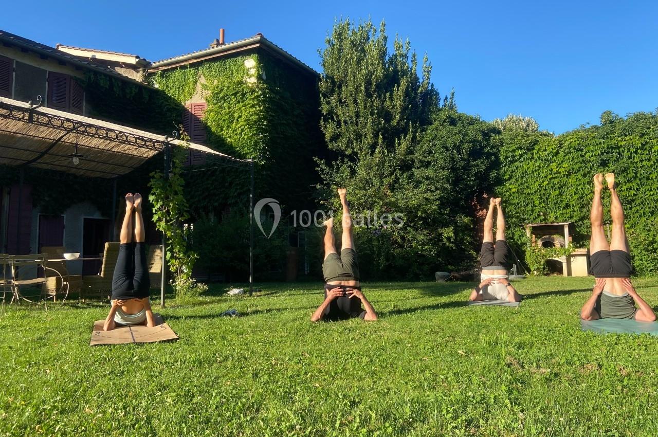 Quatre personnes pratiquent une posture de yoga inversée sur un gazon, entourées de végétation et de bâtiments couverts de…