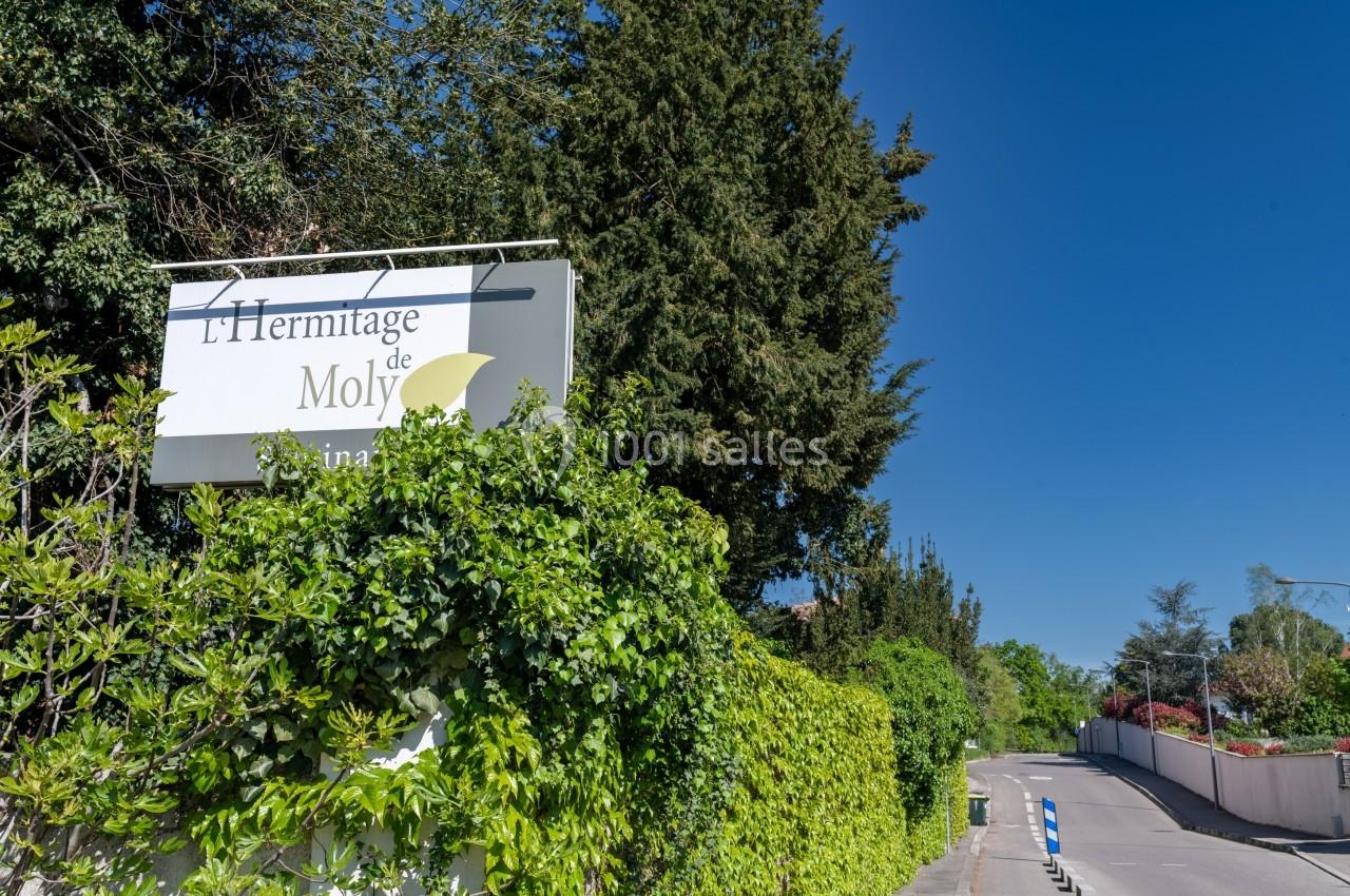 Panneau de l'Hermitage de Moly entouré de végétation, situé le long d'une route bordée d'arbres et de haies.