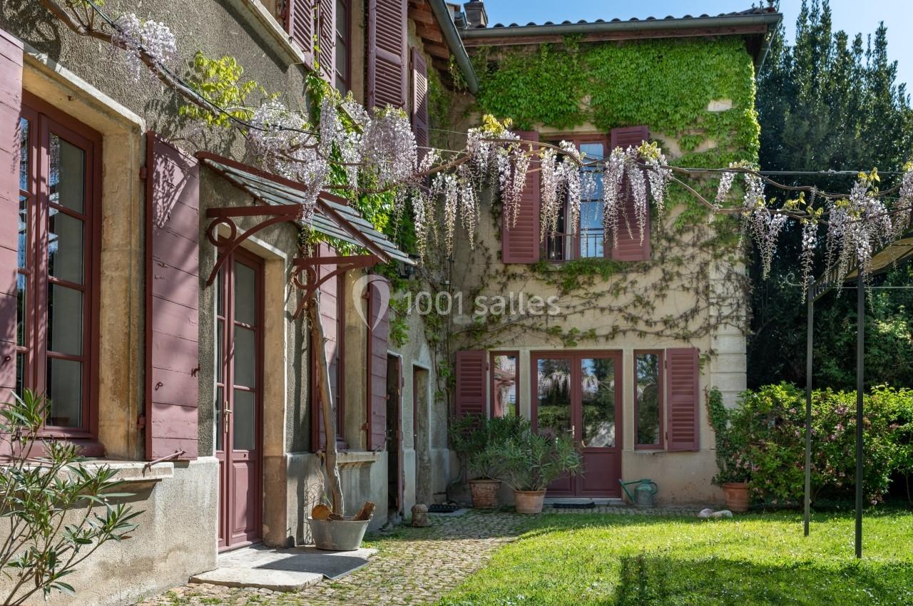Façade d'une maison ancienne avec volets rouges, glycines en fleurs et jardin verdoyant.
