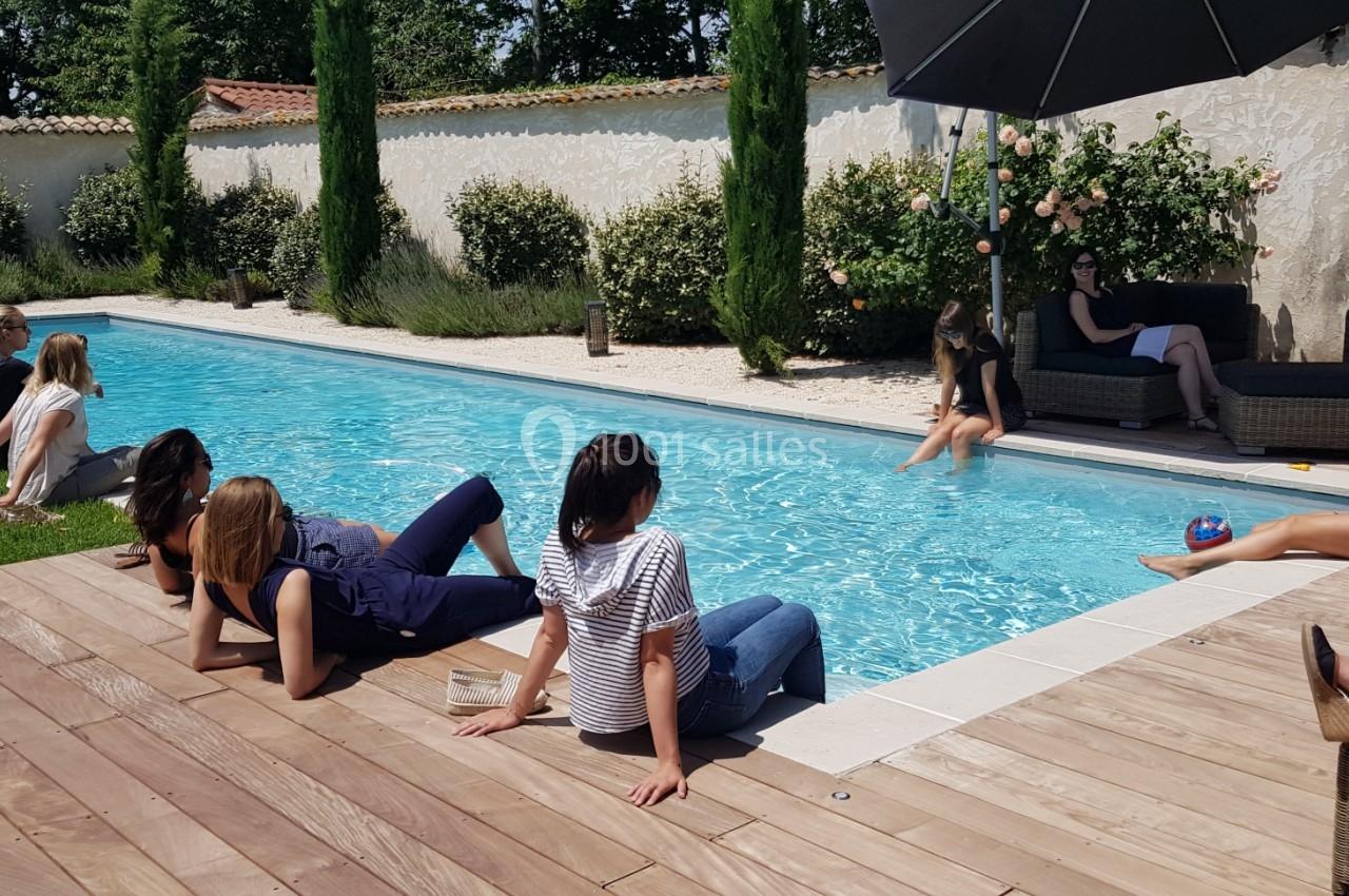 Des personnes assises ou allongées près d'une piscine dans un jardin, certaines discutant ou se relaxant.