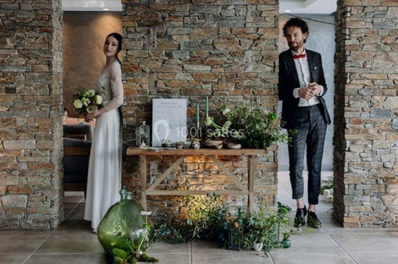 Un couple en tenue de mariage pose près d'une table décorée de plantes et d'objets dans un intérieur en pierre.