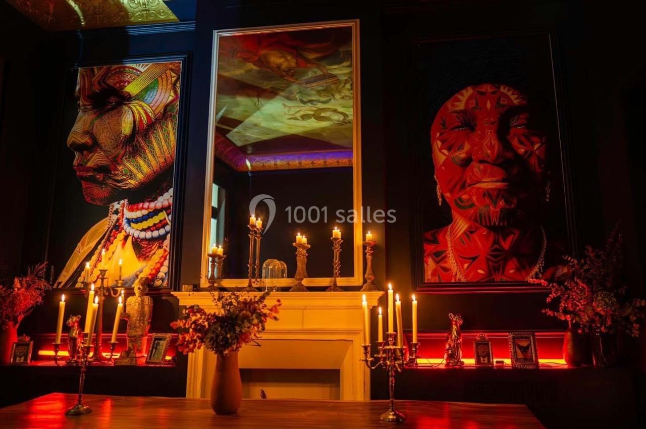 Deux portraits colorés encadrés décorent un mur sombre, éclairés par des chandeliers et des bougies sur une cheminée.