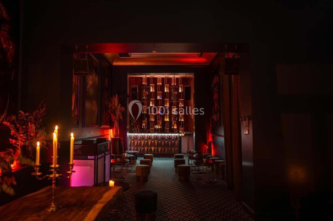 Salle sombre avec éclairage tamisé rouge, mobilier élégant, chandeliers et étagères illuminées derrière un bar.